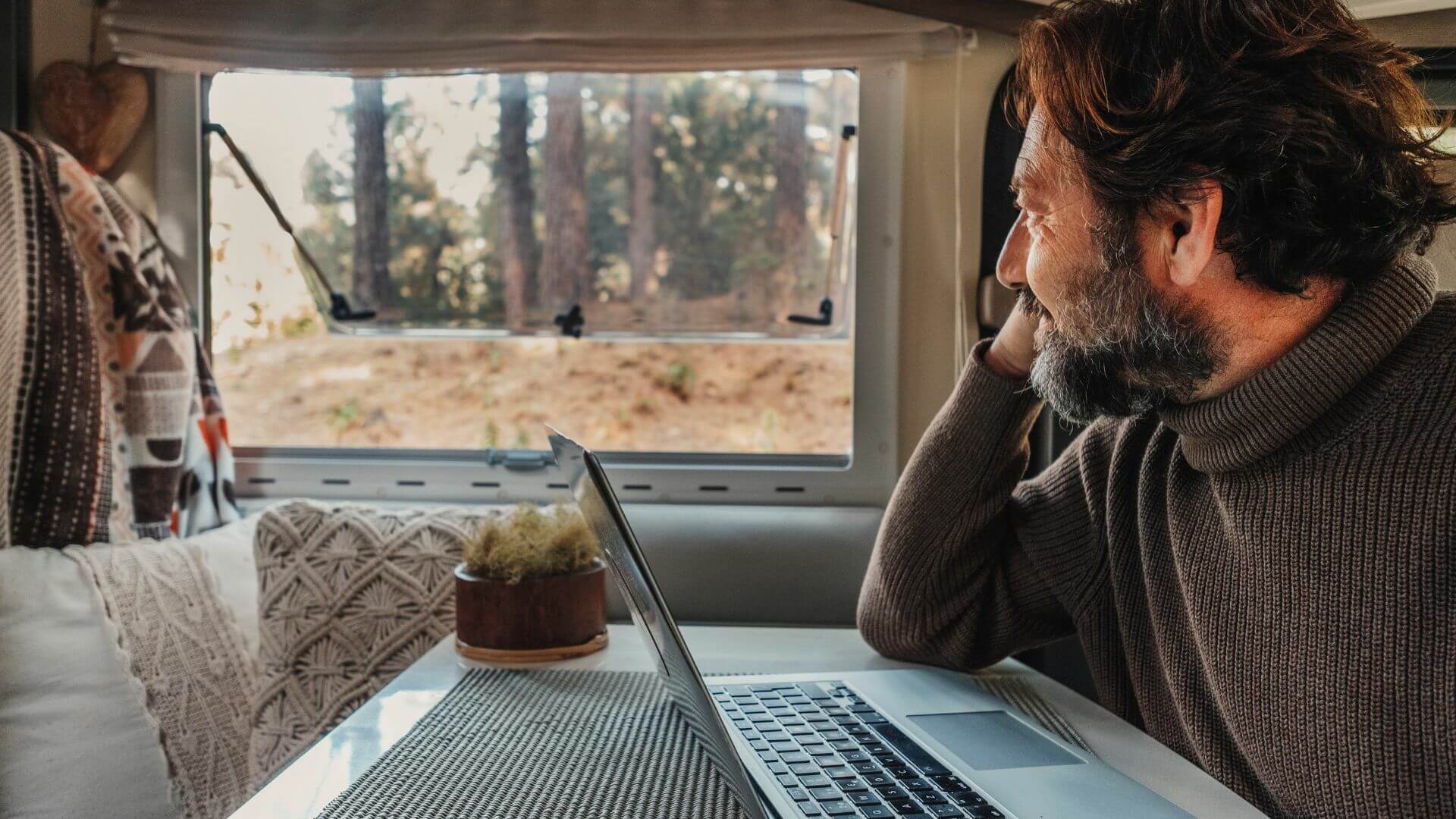 A man in a cozy setting working on a laptop while gazing out at a forested view, illustrating the flexible remote lifestyle possible with off-grid hunting properties in Michigan.

